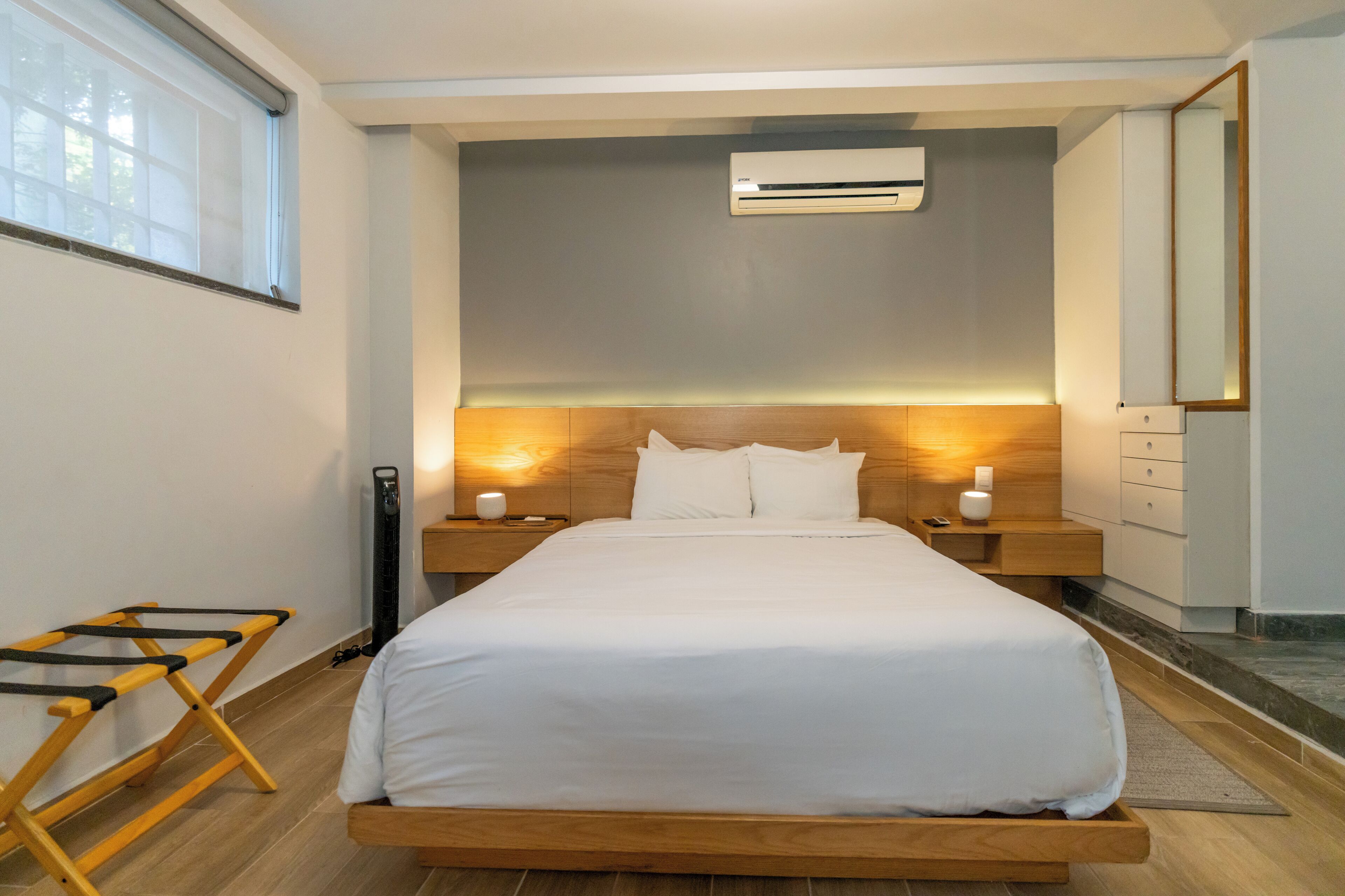 Una habitación de hotel moderna con una cama grande, cabecero de madera, mesitas de noche con lámparas, un equipo de aire acondicionado y una ventana con persianas.