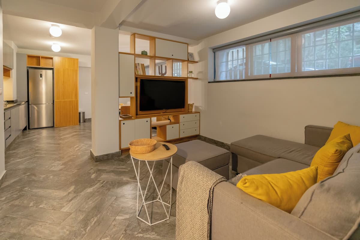 Un espacio habitable compacto con kitchenette, un sofá con cojines amarillos y un televisor sobre un soporte de madera.