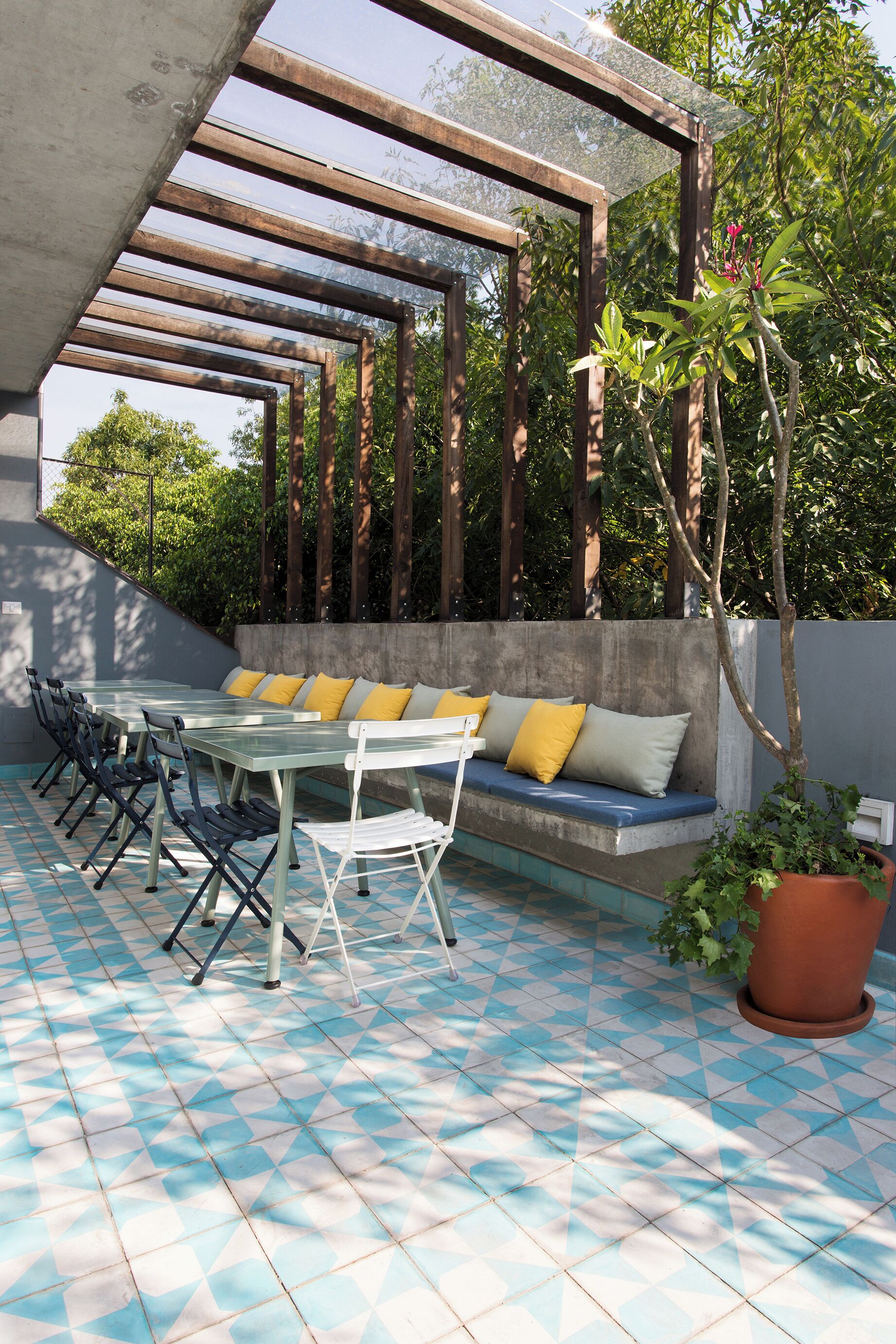 Un patio al aire libre con piso de baldosas azules y blancas, una mesa blanca con cojines amarillos y un banco de concreto con cojines.