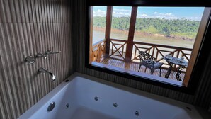 Un baño moderno con una bañera grande, una terraza de madera con mesa y sillas, y vistas a un río y un bosque.