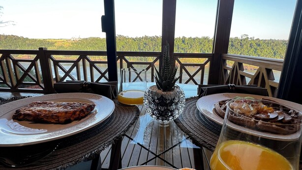 Una mesa de comedor con platos de comida, una bandeja de frutas y un vaso de jugo de naranja, frente a una vista panorámica.