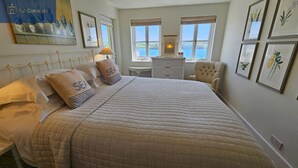 Un dormitorio con una cama grande, dos almohadas con el texto 'SEA', una mesita de noche, una silla e impresiones botánicas enmarcadas en la pared.