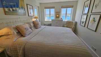 A bedroom with a large bed, two pillows with 'SEA' text, a bedside table, a chair, and framed botanical prints on the wall.