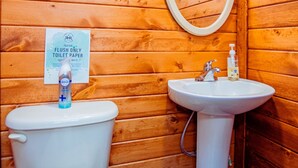 Une salle de bain rustique, aménagée dans une cabane en bois, avec des toilettes et un lavabo. Une pancarte sur le mur indique qu’il ne faut jeter dans les toilettes que du papier hygiénique.