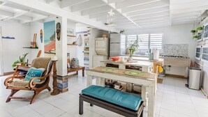 Un vaste espace de vie comprenant un fauteuil à bascule en bois, un banc, une table à manger avec un tapis vert, et un coin cuisine équipé d’une cuisinière et d’un réfrigérateur.