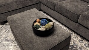 A gray sectional sofa with a black ottoman in front, a patterned rug, and a bowl of yarn on the ottoman. - New 5- Star Large, Private House, With No Neighbors  (Salina)