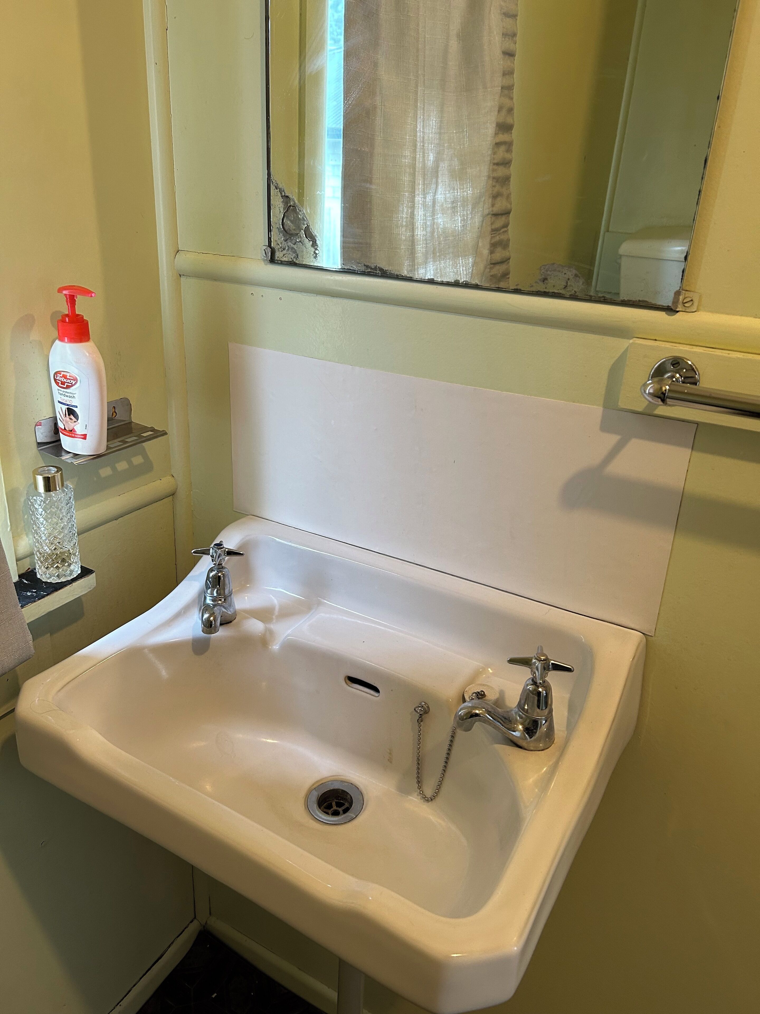 A bathroom with a white sink, two faucets, a mirror above the sink, and a bottle of lotion on a shelf.