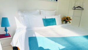 A neatly made bed with white bedding and a blue accent pillow, a blue lamp on a bedside table, and a wall-mounted coat rack with a hanger. - Haven to Relax (Hamilton)