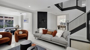 A modern living room with a grey sofa, two brown armchairs, a coffee table, and a black side table. A staircase with black railings leads to an upper level. - SuCasa Palmas (Miami)