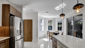A modern kitchen with a stainless steel refrigerator, wooden cabinets, a white countertop, and a dining area with a table and chairs. - SuCasa Palmas (Miami)