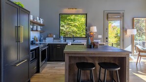 A modern kitchen with dark cabinetry, a wooden island, and a large window with a view of trees. - Big Bear Escape (Banner Elk)