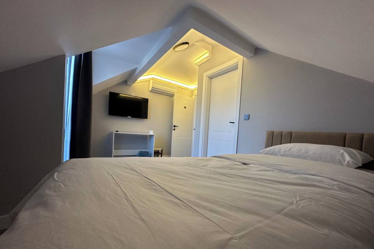 Un dormitorio con techo inclinado, una cama, un televisor montado en la pared y una puerta que da a otra habitación.