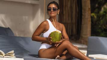 Una persona sentada en una silla reclinable azul sobre una playa arenosa, sosteniendo un coco verde.