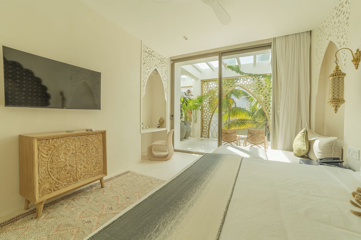 Un dormitorio moderno con una cama grande, una consola de madera, un televisor y un balcón con vistas a palmeras.