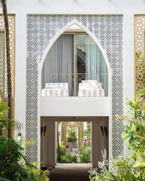 Relax on the balcony overlooking a lush garden with a serene view, featuring a cushioned seating area and arched doorway with intricate tile patterns.