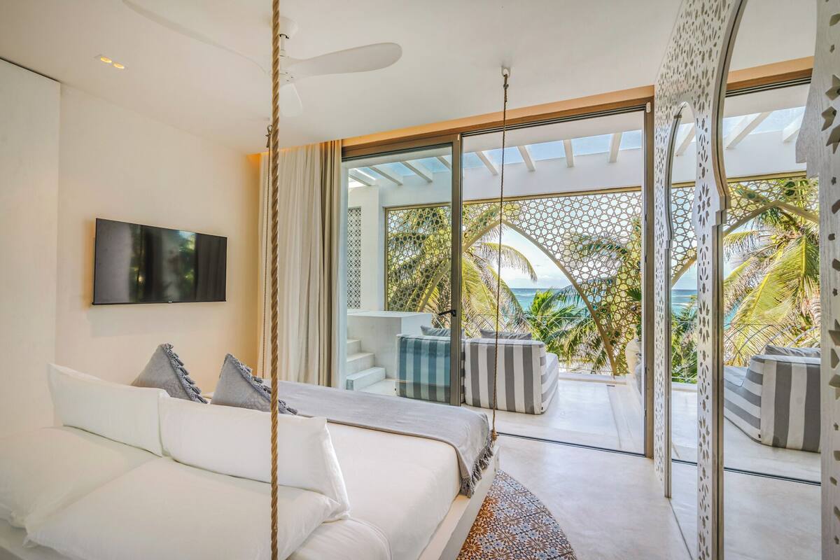 Un dormitorio moderno con una cama grande, un televisor montado en la pared y un balcón con vistas a palmeras y al océano.