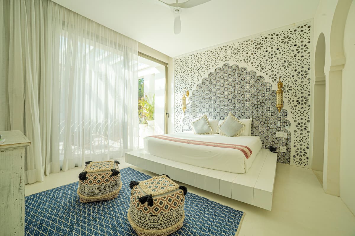 Un dormitorio con cama, un diseño decorativo en la pared, una ventana con cortinas y dos otomanas.