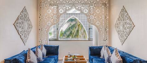 A room with two blue sofas, a wooden coffee table, and a patterned ottoman. A decorative wall with intricate cutouts and a large window with a view of palm trees.