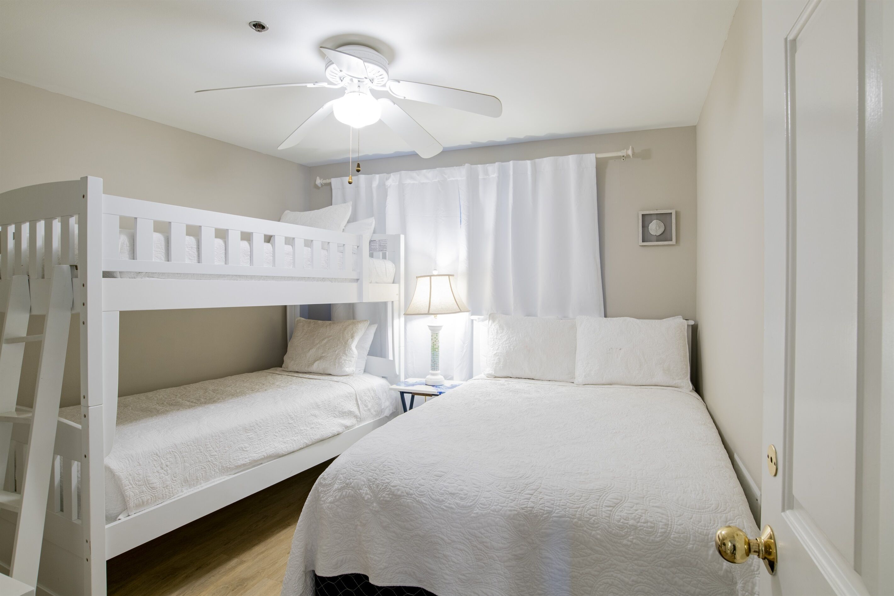 Une chambre avec deux lits superposés, un ventilateur de plafond, une table de chevet avec une lampe et une fenêtre avec des rideaux.