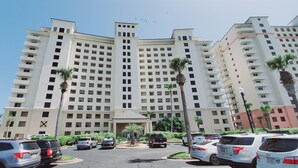 A multi-story hotel with a central entrance, surrounded by parked cars and palm trees.