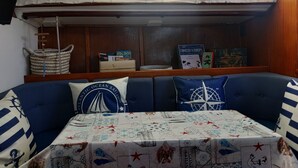 A cabin with a blue sofa, a table with a nautical-themed tablecloth, and books on a shelf. - Alojamento Barco Casa Ancorado no Coração da Ria Formosa em Faro (Faro)