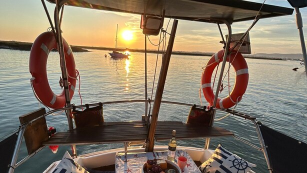 A boat deck with a table set for dining, life rings, and a view of the sunset over the water. - Alojamento Barco Casa Ancorado no Coração da Ria Formosa em Faro (Faro)