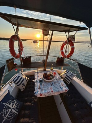 Un pont de bateau avec une table dressée pour le dîner, des bouées de sauvetage et une vue sur le coucher de soleil sur l’eau.