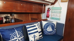 A cabin with a wooden bench, nautical-themed pillows, and a wall-mounted sign saying "There is no place like the beach." - Alojamento Barco Casa Ancorado no Coração da Ria Formosa em Faro (Faro)