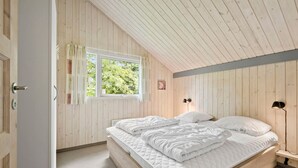 Ein Schlafzimmer mit schrägen Wänden, Holzwänden, einem Fenster mit Vorhängen, einem Bett mit weißer Bettwäsche und einem Nachttisch mit einer Lampe.