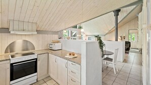 Eine moderne Küche mit weißen Schränken, einem Edelstahlbackofen und einer Mikrowelle. Es gibt einen Essbereich mit Tisch und Stühlen sowie einen Blick ins Freie durch große Fenster.