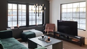 A modern living room with a large flat-screen TV, a green sofa, a coffee table with books and candles, and a fireplace.