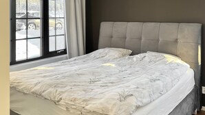 A bed with a grey upholstered headboard, a white bedspread, and a window with a view of parked cars.