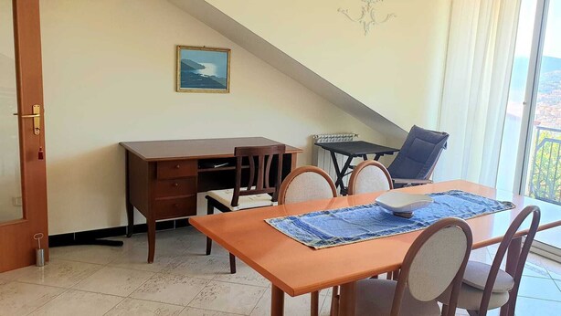 Un coin repas comprenant une table, des chaises, un petit bureau et un fauteuil. Une fenêtre offre une vue sur le paysage urbain.