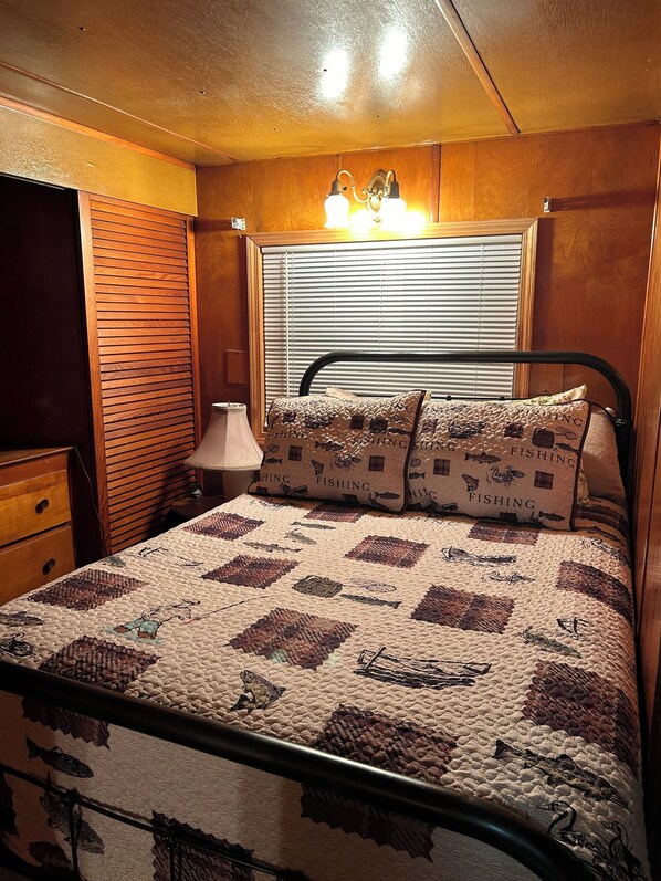 Una cama con colcha decorada con motivos de peces y pesca, una lámpara de noche y una ventana con persianas.