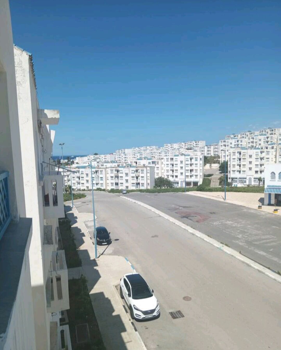 A residential area with white apartment buildings, a clear blue sky, and a road with parked cars.