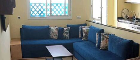 A living room with a blue sofa, a small table, and a kitchen area visible through an open door.