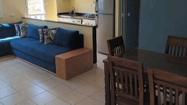 Een woonkamer met een blauwe bank, een houten eettafel en stoelen. Er is een keuken met houten keukenkastjes en een koelkast.
