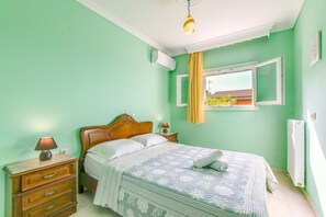Ein Schlafzimmer mit einem Bett, einem Nachttisch, einer Lampe, einem Fenster mit Vorhängen und einer Klimaanlage.