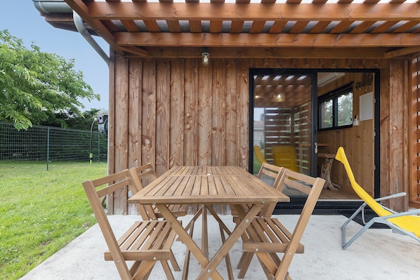 Un'area pranzo esterna in legno con tavolo e sedie, una poltrona da relax gialla e un edificio in legno con tetto spiovente.