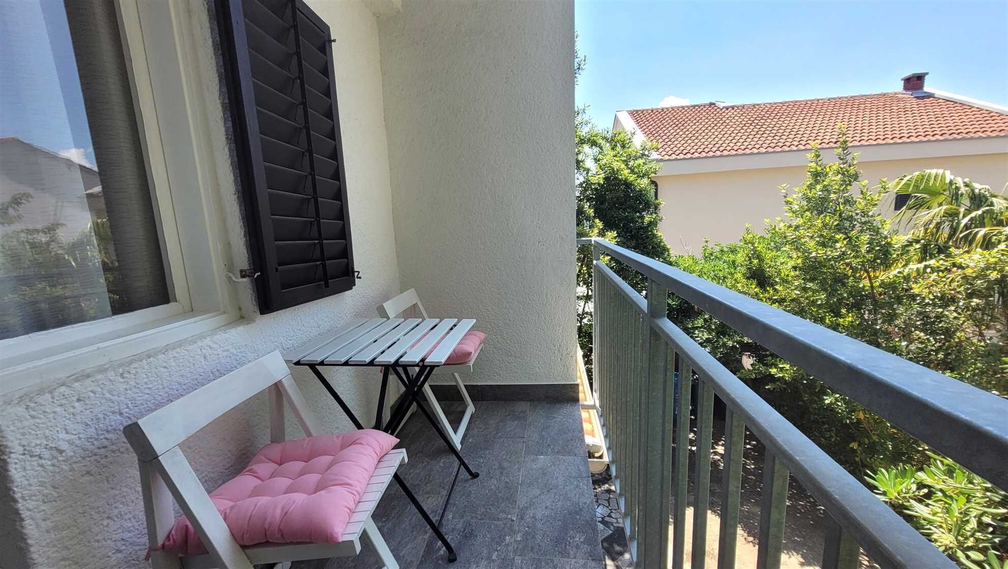 A balcony with a white chair and a small table, a window with black shutters, and a view of a building with a red roof.