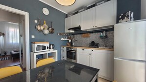 A compact kitchen with white cabinets, a black countertop, and a microwave. There is a clock on the wall and a window with curtains. - Apartment Lončar with sea view (Krk)