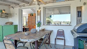A rustic outdoor dining area with a wooden table and metal chairs, a ceiling fan, a large window with a view, and a barbecue grill.