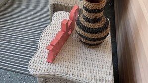 Ein Korbsessel mit Kissen, ein kleiner Tisch und eine dekorative Vase auf einem Balkon mit Blick auf ein Gebäude.