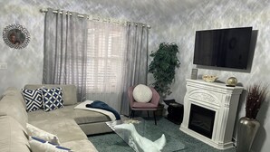 Ein Wohnzimmer mit einem beigefarbenen Sofa, gemusterten Kissen, einem Glastisch, einem weißen Kamin, einem Fernseher und einer großen Pflanze.