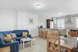 Een moderne woonkamer met een blauwe bank, een witte salontafel en houten stoelen. In de achtergrond is een keuken met een magnetron en een groot raam zichtbaar.