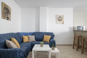 Een woonkamer met een blauwe bank, een witte salontafel en een klein keukentje met een magnetron.