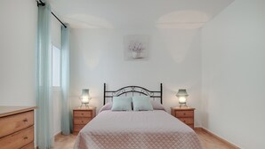 Een slaapkamer met een bed, twee nachtkastjes met lampen, een houten dressoir, een raam met gordijnen en een aan de muur bevestigd schilderij.