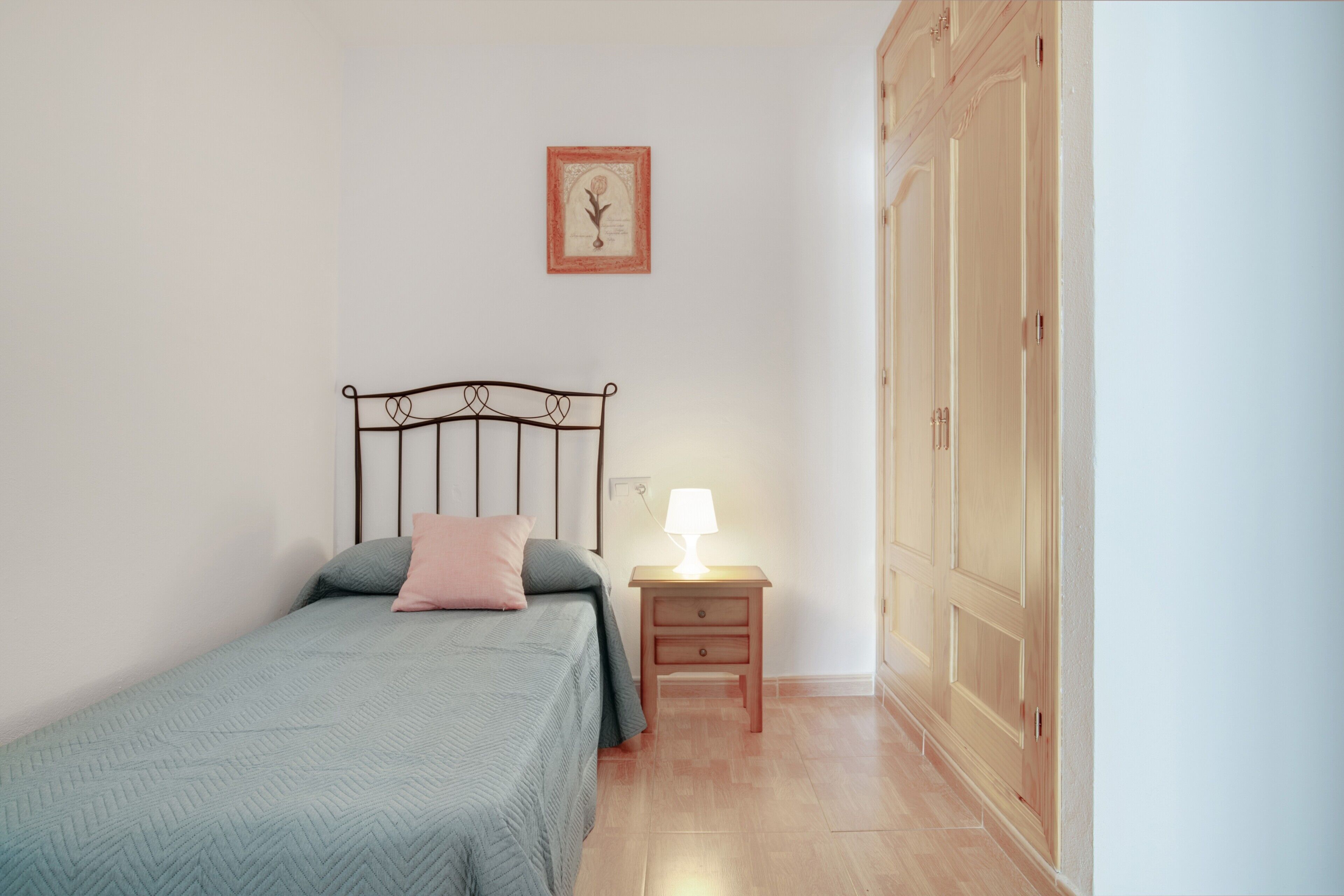 A bedroom with a bed, bedside table, lamp, wardrobe, and a framed picture on the wall.