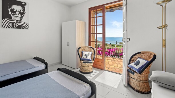 A bedroom with a bed, a wardrobe, a wicker chair, a bedside table, and a view of the outside. - Holiday Home 'La Belle Provence' with Sea View, Wi-Fi and Air Conditioning (Roquebrune-sur-Argens)