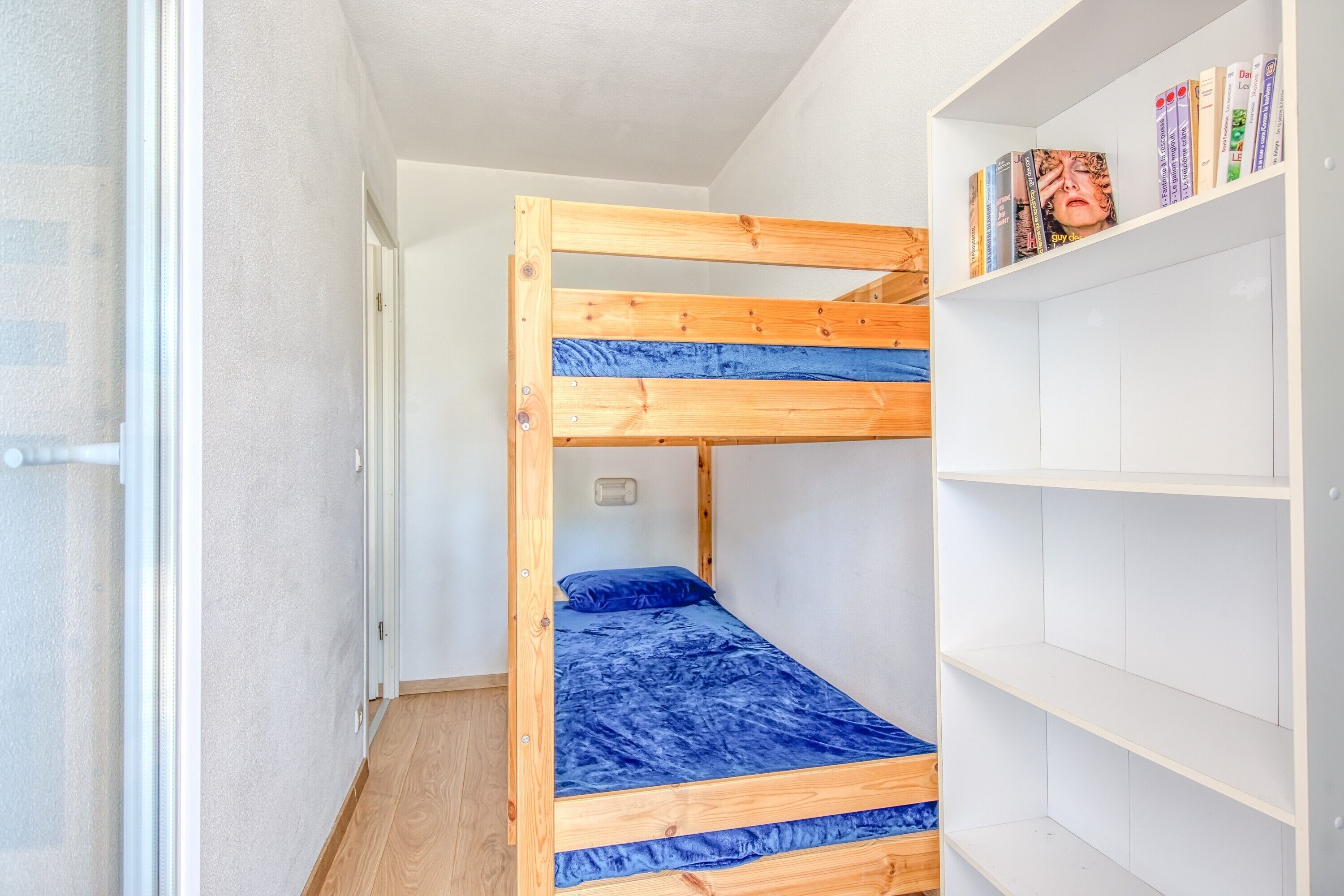 A bunk bed with a blue mattress, a white bookshelf with books, and a wooden door.